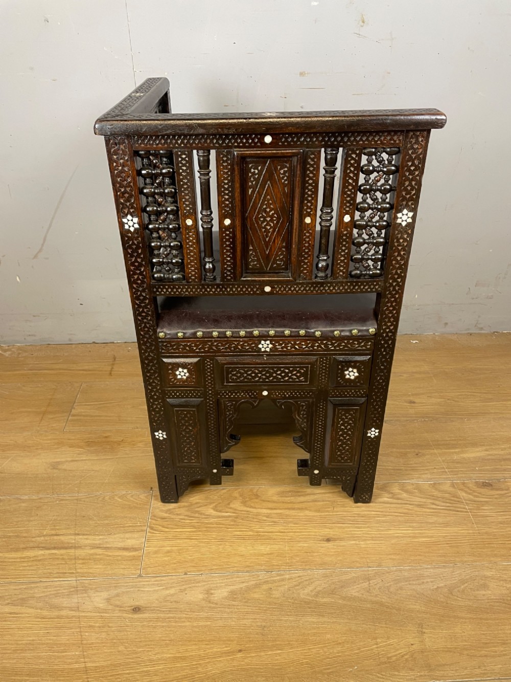 A 19th-century carved dark wood corner chair featuring mother-of-pearl floral inlay, detailed Mashrabiya panels, and a brown leather seat secured with brass studs. View VI