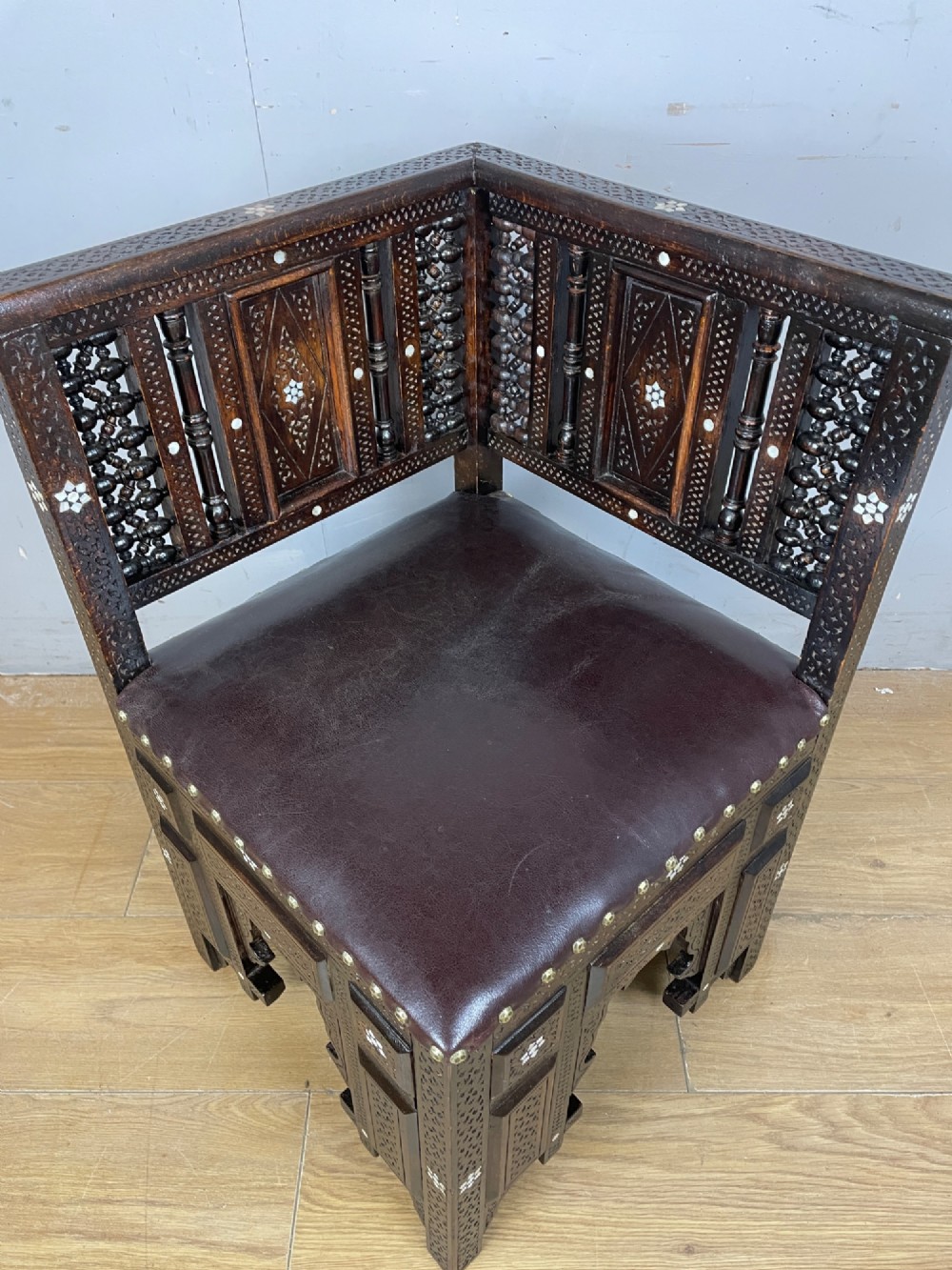 A 19th-century carved dark wood corner chair featuring mother-of-pearl floral inlay, detailed Mashrabiya panels, and a brown leather seat secured with brass studs. View VII