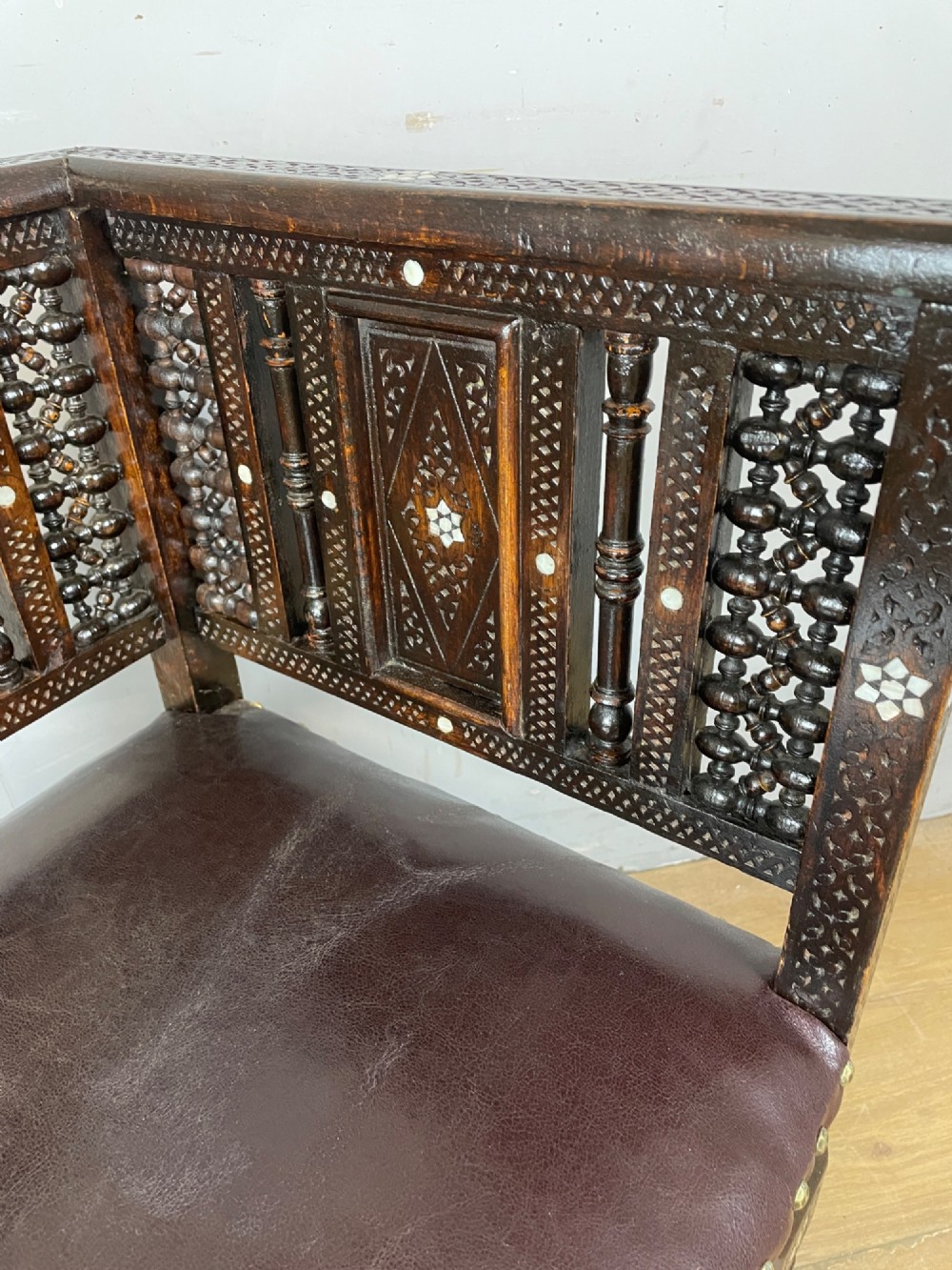 A 19th-century carved dark wood corner chair featuring mother-of-pearl floral inlay, detailed Mashrabiya panels, and a brown leather seat secured with brass studs. View X
