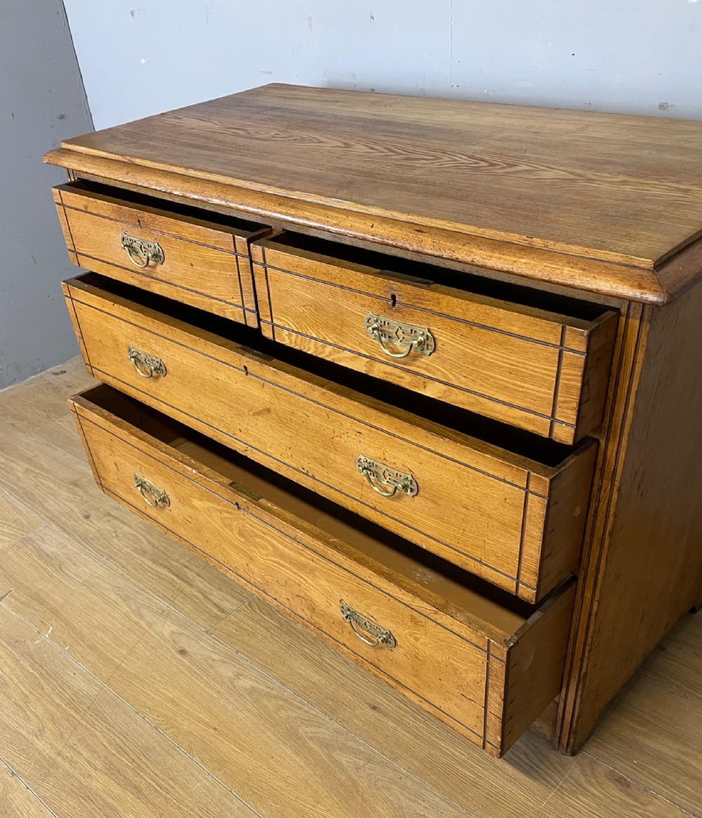 An Arts and Crafts solid ash chest of drawers featuring two short drawers over two long drawers with brass drop handles and dark linear inlay. View V