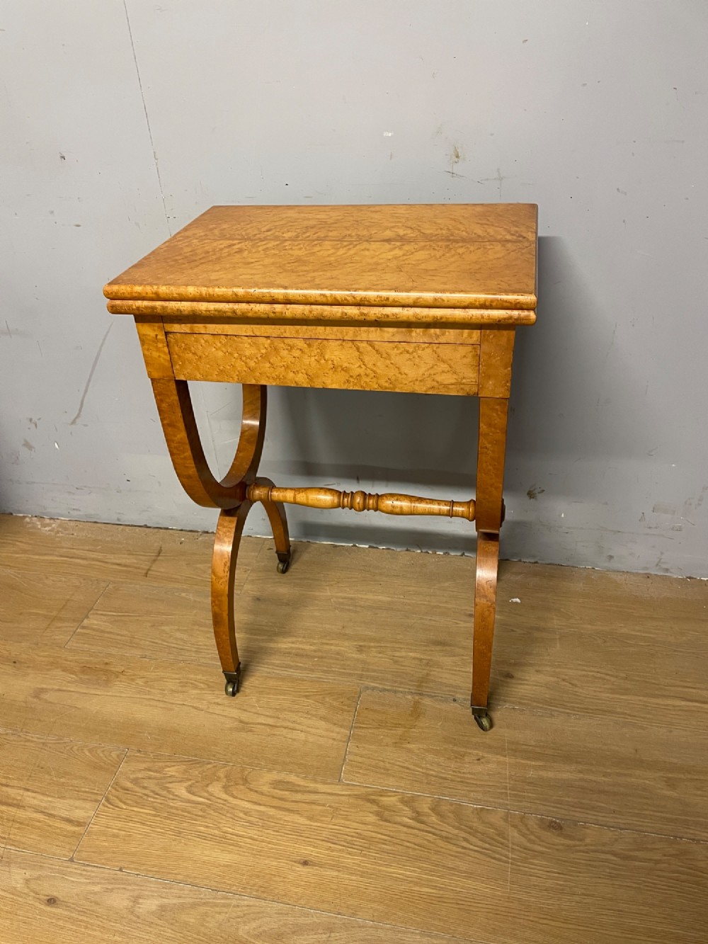 A 19th-century Biedermeier birds eye maple card table featuring a rectangular fold-over top, sweeping legs, a turned central stretcher, and brass castors.