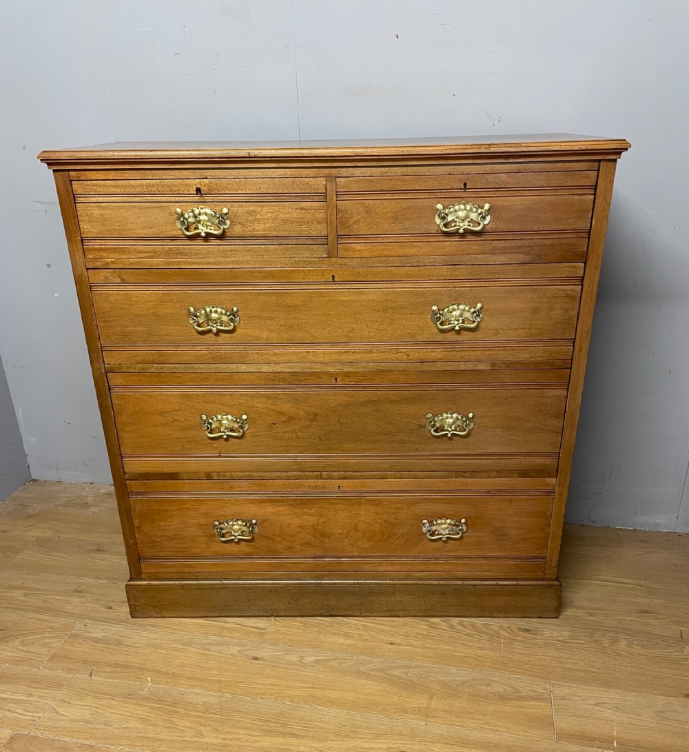 An Arts and Crafts satin walnut chest of drawers featuring two short over three long drawers with brass drop handles and a solid plinth base.