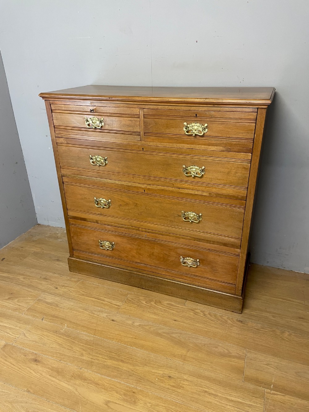 An Arts and Crafts satin walnut chest of drawers featuring two short over three long drawers with brass drop handles and a solid plinth base. View III