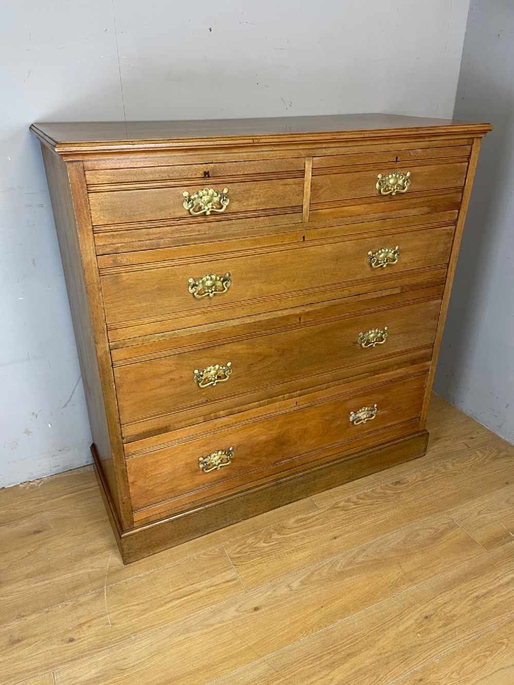 An Arts and Crafts satin walnut chest of drawers featuring two short over three long drawers with brass drop handles and a solid plinth base. View XI