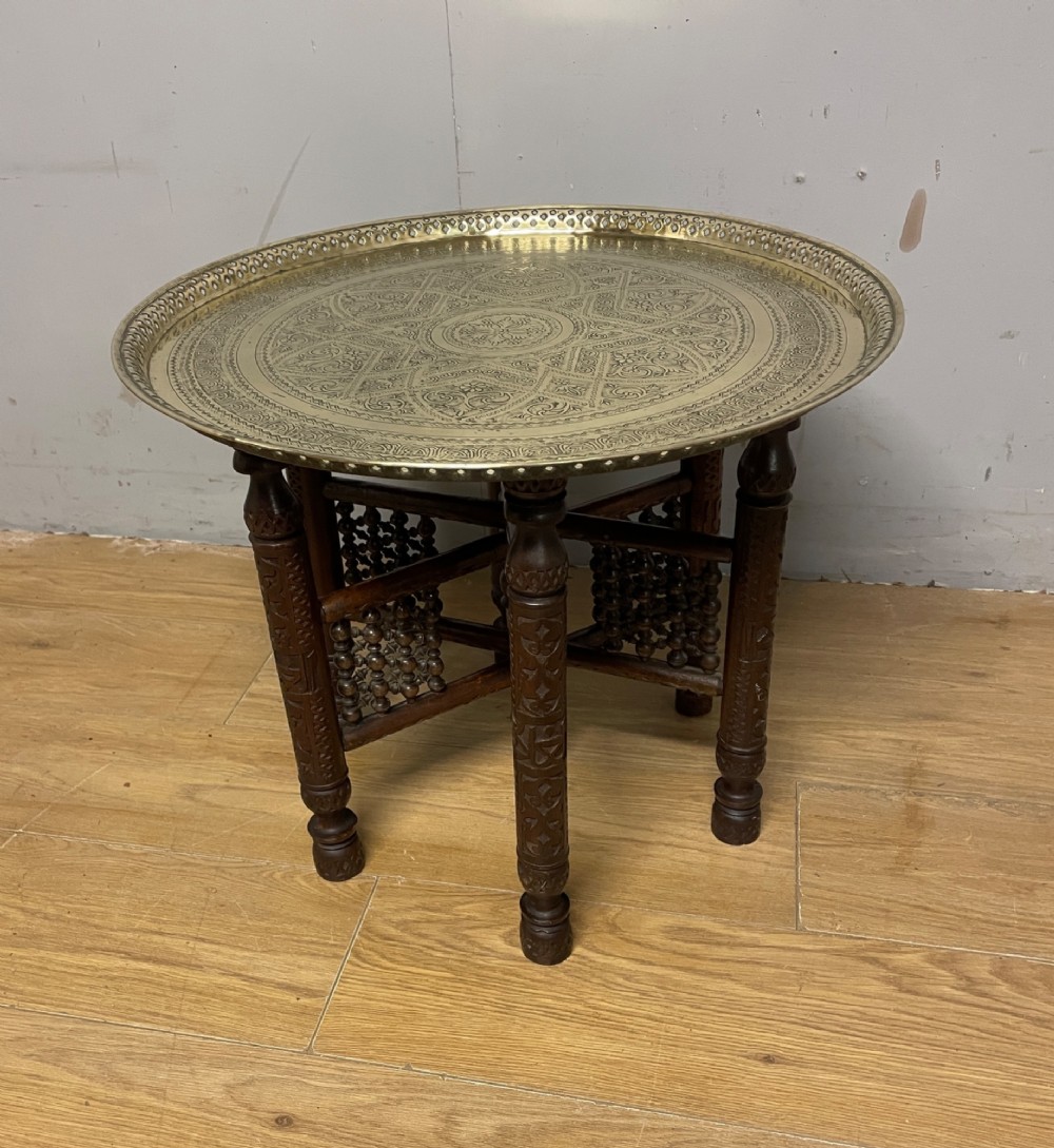 A late 19th-century Anglo-Indian Benares table featuring an intricately engraved circular brass tray top resting on a dark carved wooden folding base.