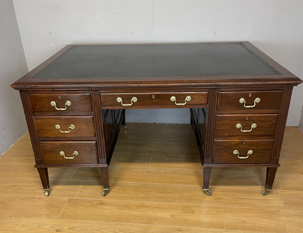 An Edwardian mahogany partners desk featuring a green leather writing surface, brass swan-neck handles, fluted pilasters, and tapered legs on castors.