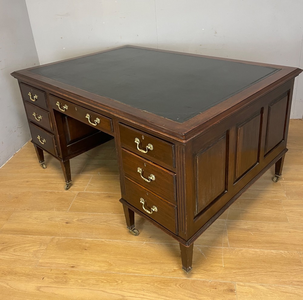 An Edwardian mahogany partners desk featuring a green leather writing surface, brass swan-neck handles, fluted pilasters, and tapered legs on castors. View III