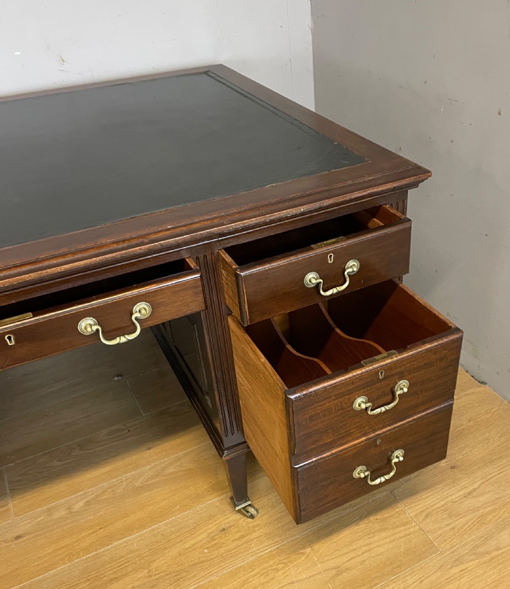 An Edwardian mahogany partners desk featuring a green leather writing surface, brass swan-neck handles, fluted pilasters, and tapered legs on castors. View IV