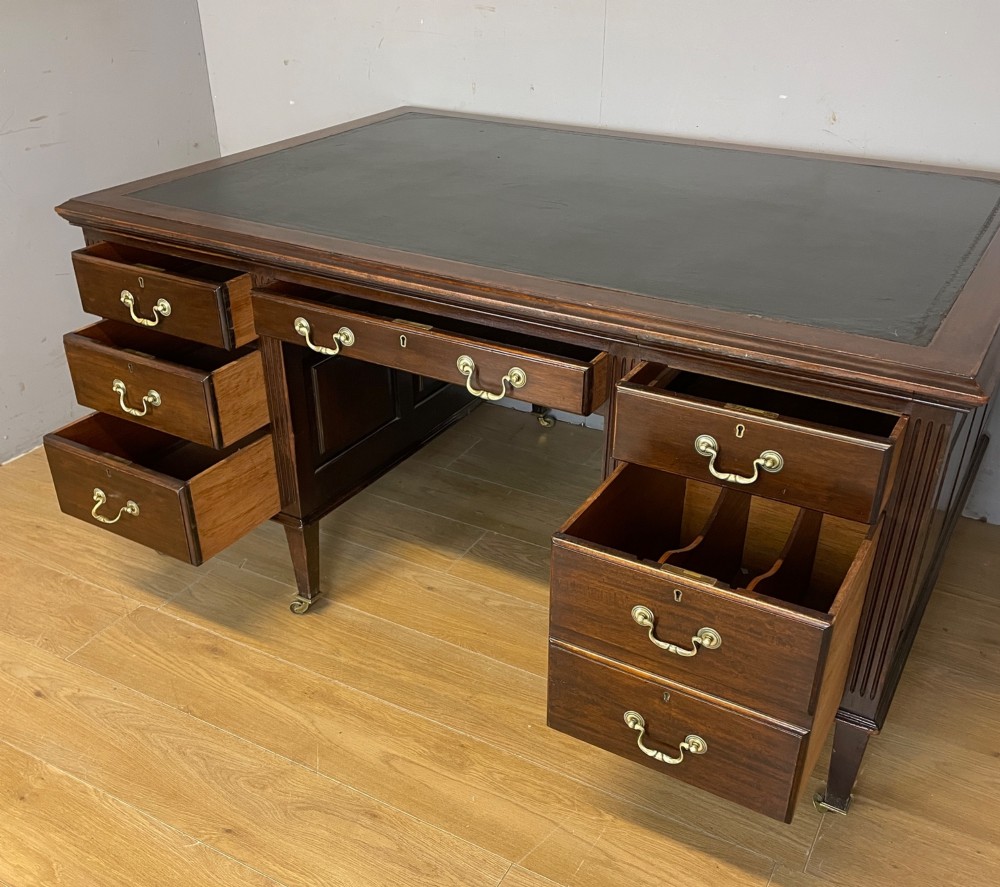 An Edwardian mahogany partners desk featuring a green leather writing surface, brass swan-neck handles, fluted pilasters, and tapered legs on castors. View V