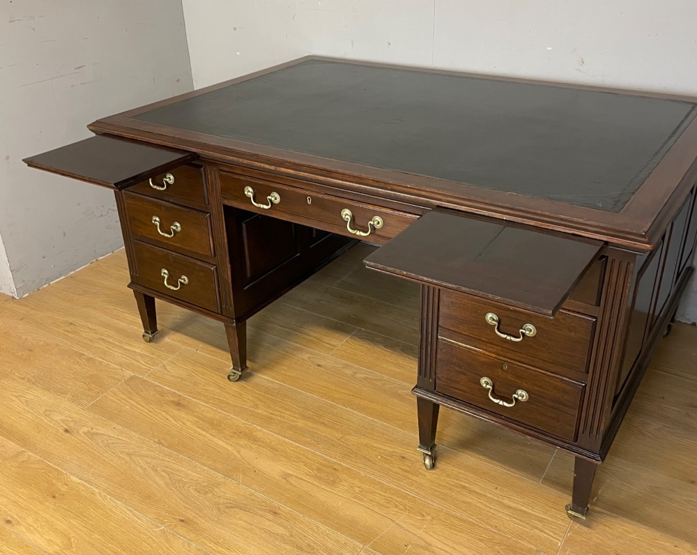 An Edwardian mahogany partners desk featuring a green leather writing surface, brass swan-neck handles, fluted pilasters, and tapered legs on castors. View VI