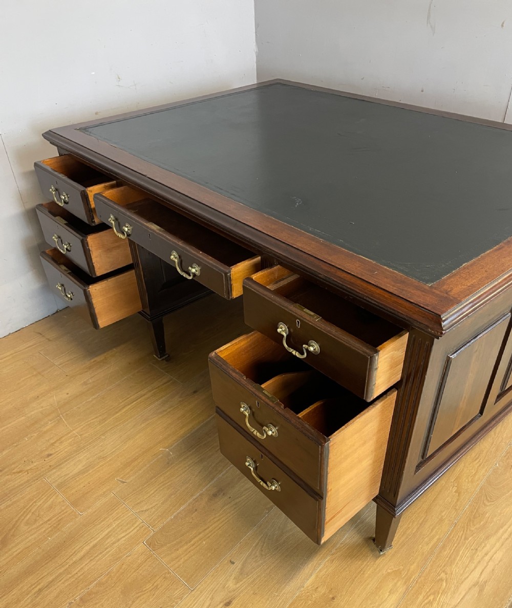 An Edwardian mahogany partners desk featuring a green leather writing surface, brass swan-neck handles, fluted pilasters, and tapered legs on castors. View IX