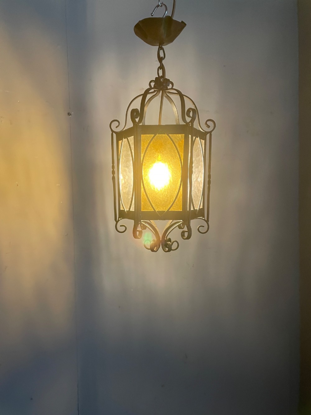 An illuminated hexagonal wrought iron hall lantern with textured amber glass panels and decorative scrollwork.