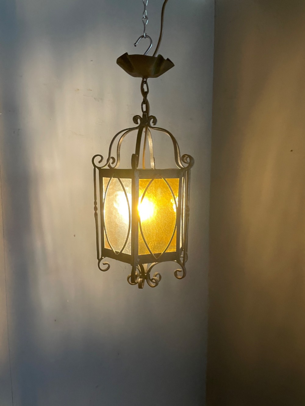 An illuminated hexagonal wrought iron hall lantern with textured amber glass panels and decorative scrollwork. View II