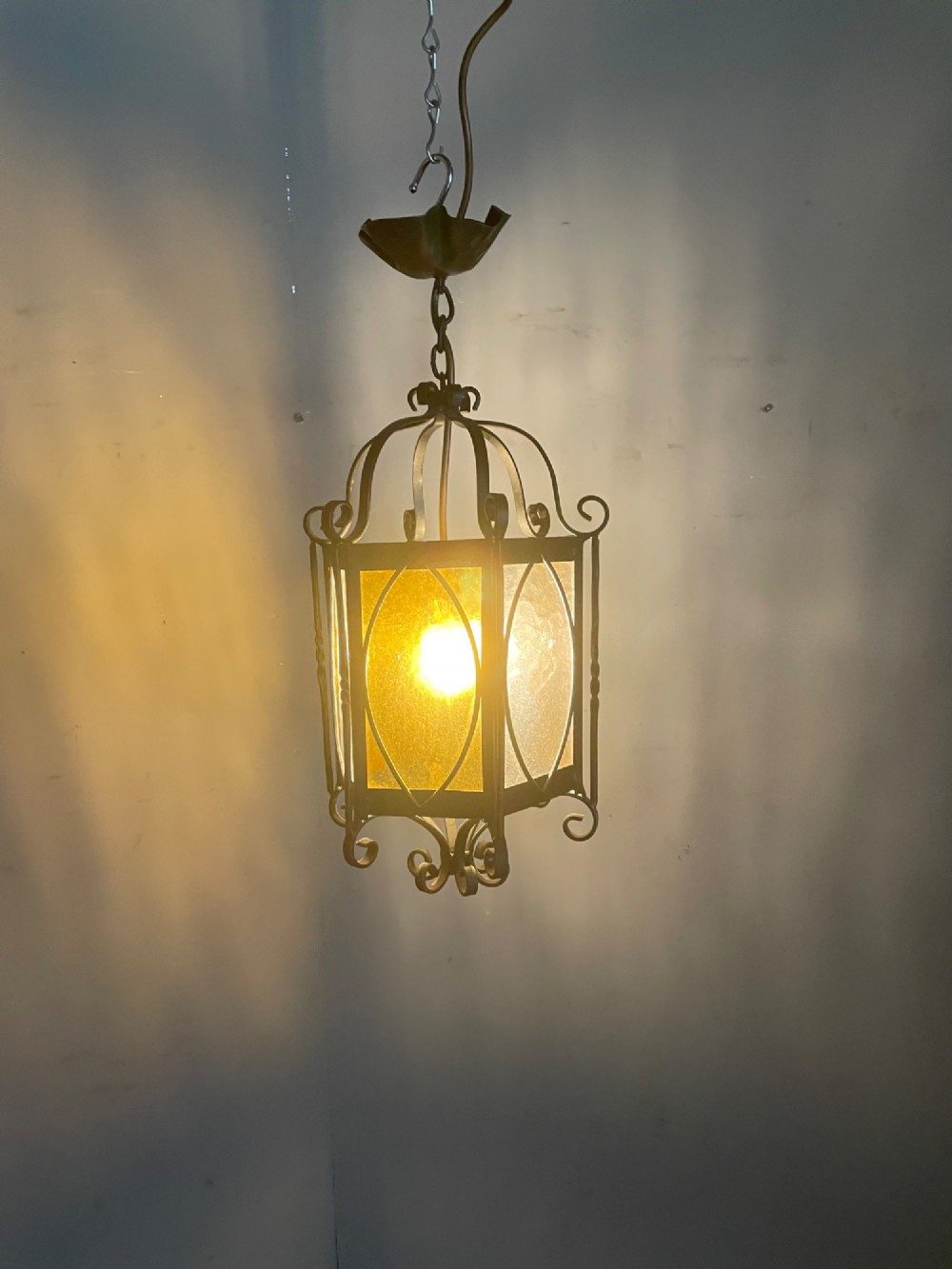 An illuminated hexagonal wrought iron hall lantern with textured amber glass panels and decorative scrollwork. View III