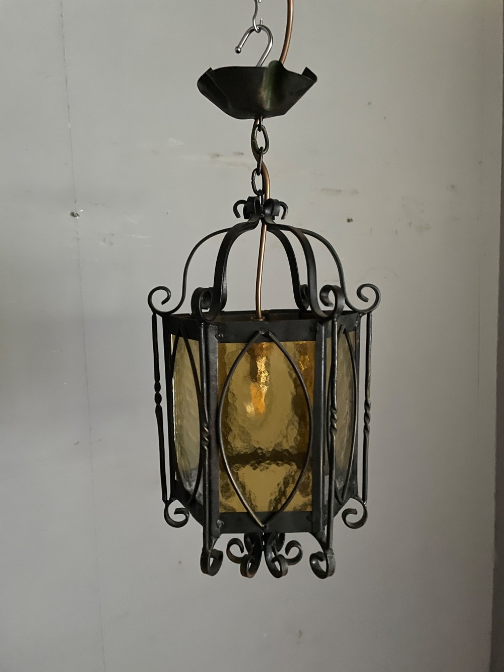 An illuminated hexagonal wrought iron hall lantern with textured amber glass panels and decorative scrollwork. View IV