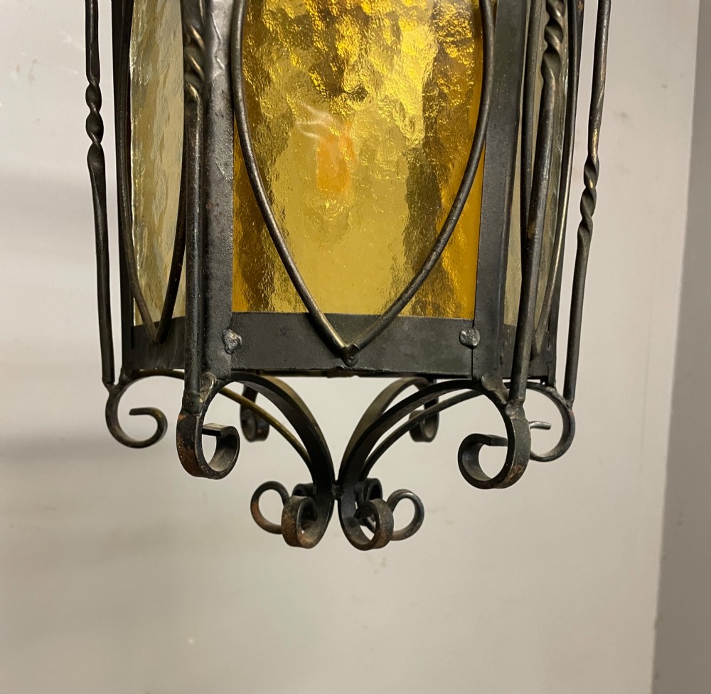 An illuminated hexagonal wrought iron hall lantern with textured amber glass panels and decorative scrollwork. View VII