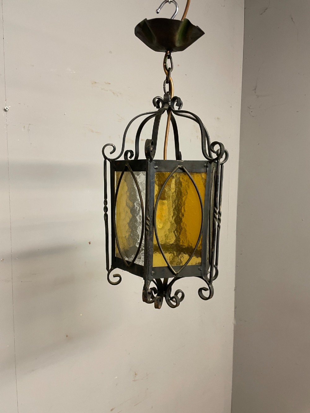 An illuminated hexagonal wrought iron hall lantern with textured amber glass panels and decorative scrollwork. View VIII