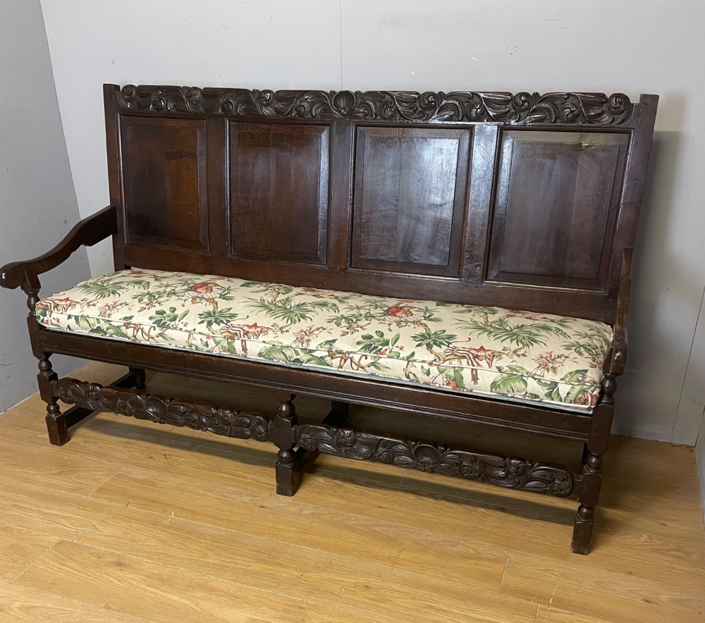 An antique dark oak wooden settle featuring four raised back panels, detailed floral relief carving along the crest and apron, turned legs, and a floral pattern cushion.
