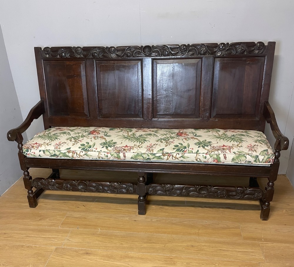 An antique dark oak wooden settle featuring four raised back panels, detailed floral relief carving along the crest and apron, turned legs, and a floral pattern cushion. View II