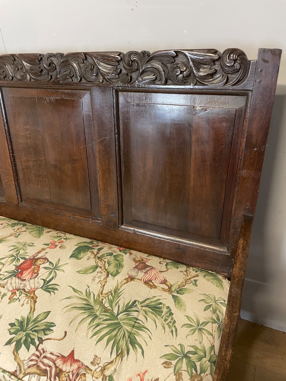 An antique dark oak wooden settle featuring four raised back panels, detailed floral relief carving along the crest and apron, turned legs, and a floral pattern cushion. View III