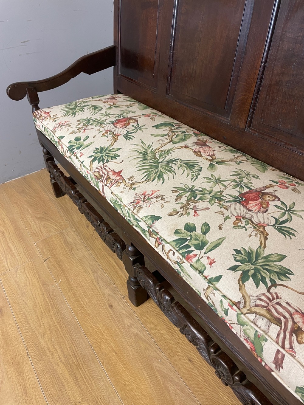 An antique dark oak wooden settle featuring four raised back panels, detailed floral relief carving along the crest and apron, turned legs, and a floral pattern cushion. View IV