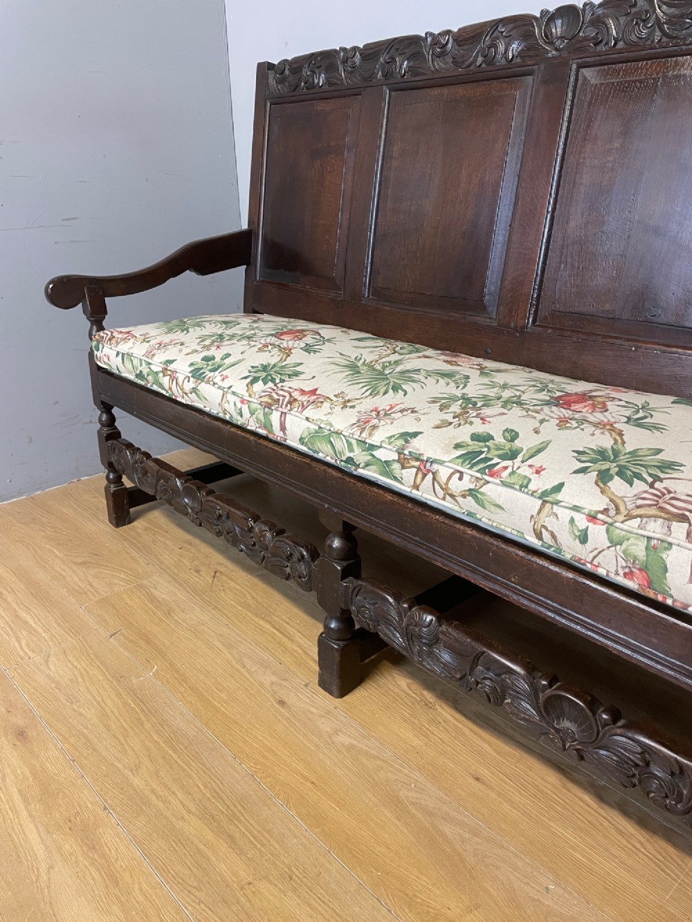 An antique dark oak wooden settle featuring four raised back panels, detailed floral relief carving along the crest and apron, turned legs, and a floral pattern cushion. View V