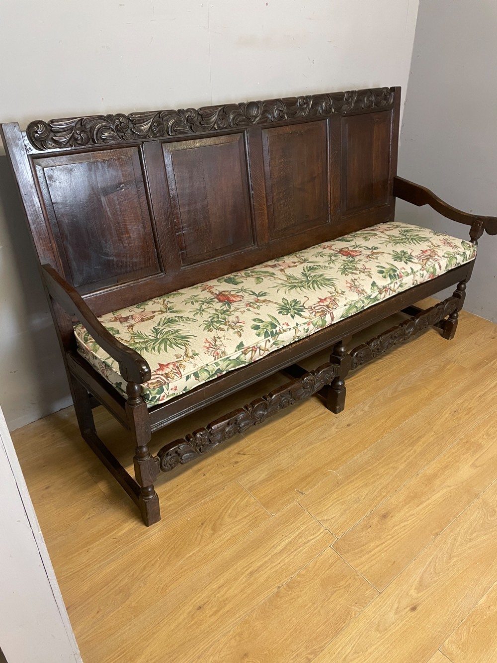 An antique dark oak wooden settle featuring four raised back panels, detailed floral relief carving along the crest and apron, turned legs, and a floral pattern cushion. View VI
