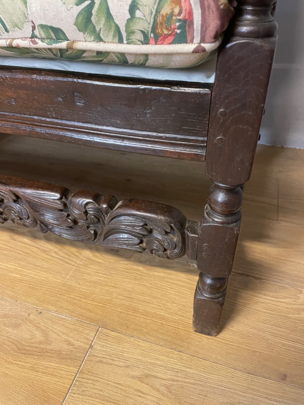 An antique dark oak wooden settle featuring four raised back panels, detailed floral relief carving along the crest and apron, turned legs, and a floral pattern cushion. View VII