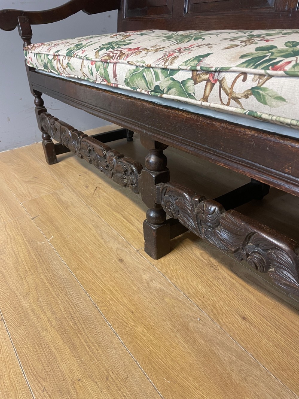 An antique dark oak wooden settle featuring four raised back panels, detailed floral relief carving along the crest and apron, turned legs, and a floral pattern cushion. View VIII