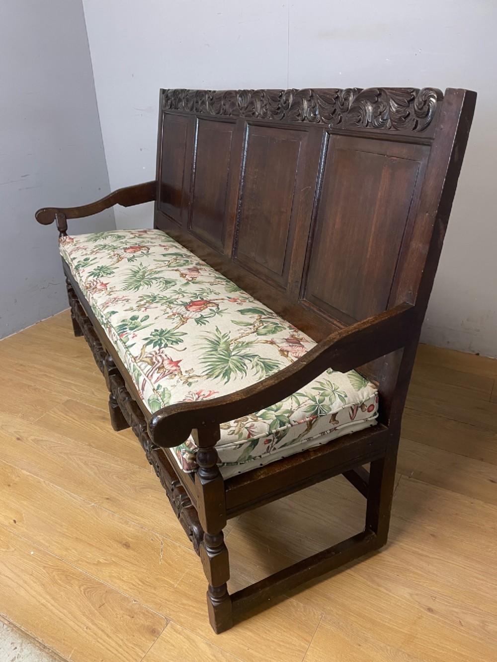 An antique dark oak wooden settle featuring four raised back panels, detailed floral relief carving along the crest and apron, turned legs, and a floral pattern cushion. View IX