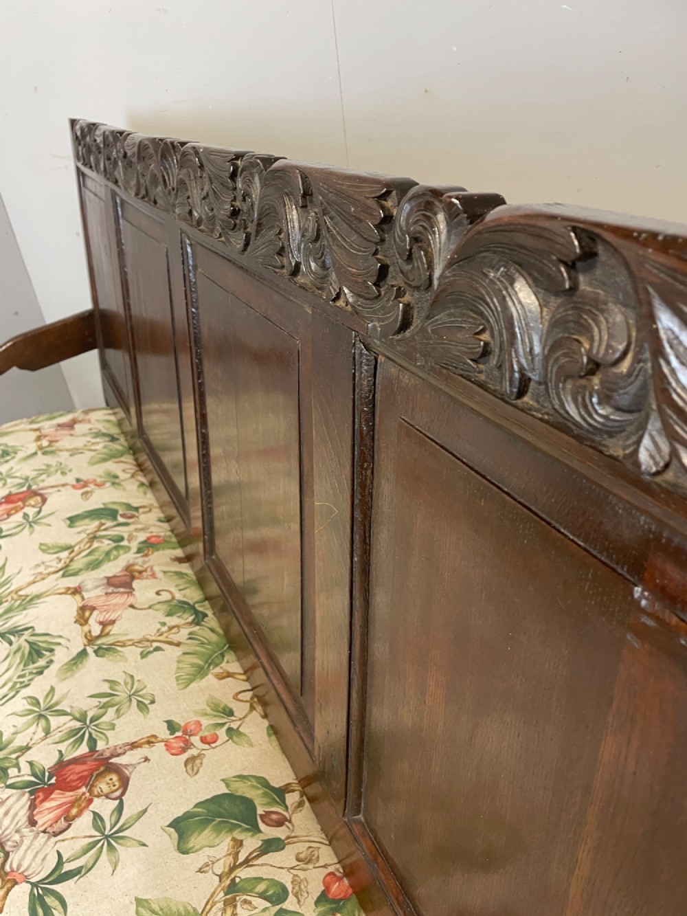 An antique dark oak wooden settle featuring four raised back panels, detailed floral relief carving along the crest and apron, turned legs, and a floral pattern cushion. View X