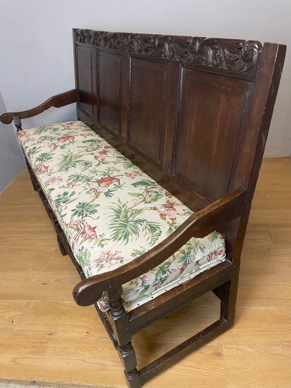 An antique dark oak wooden settle featuring four raised back panels, detailed floral relief carving along the crest and apron, turned legs, and a floral pattern cushion. View XI