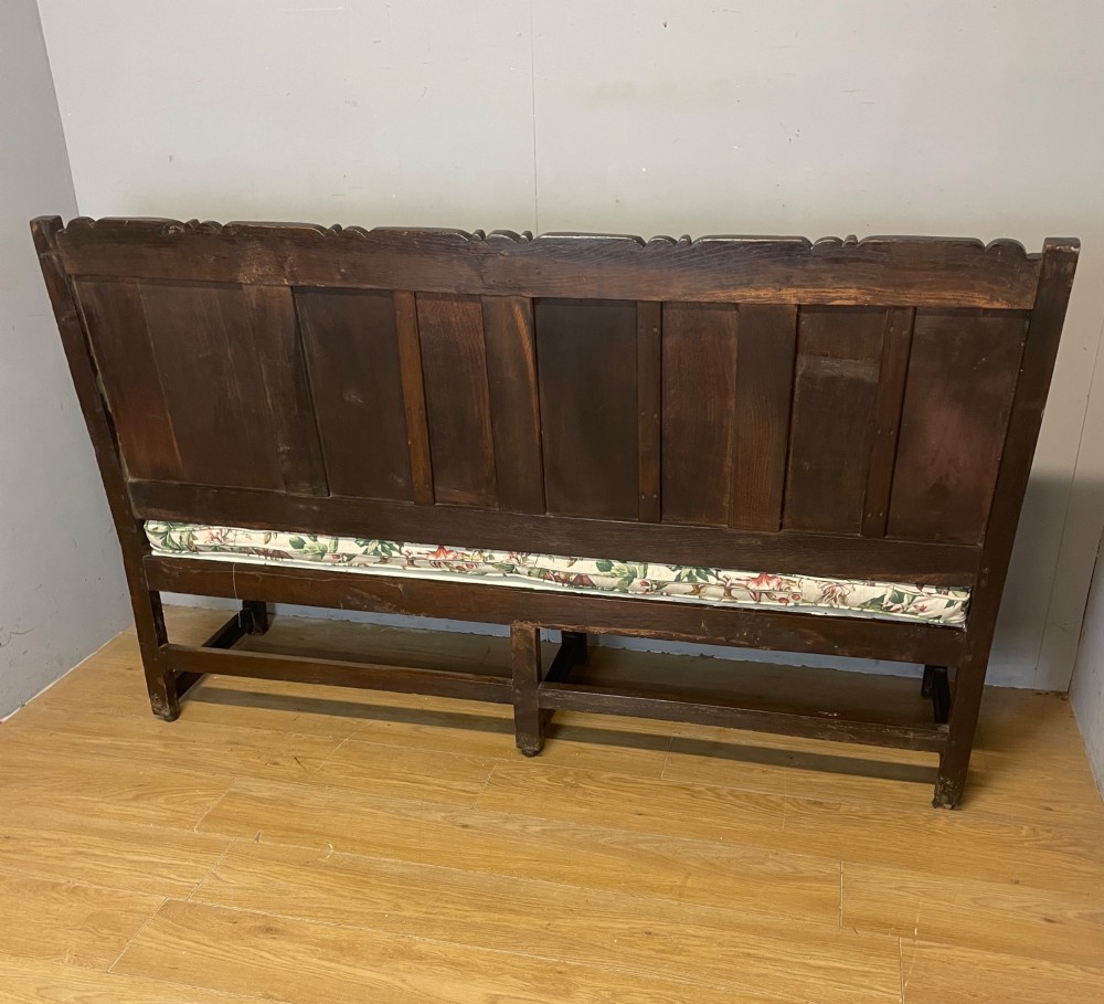 An antique dark oak wooden settle featuring four raised back panels, detailed floral relief carving along the crest and apron, turned legs, and a floral pattern cushion. View XII