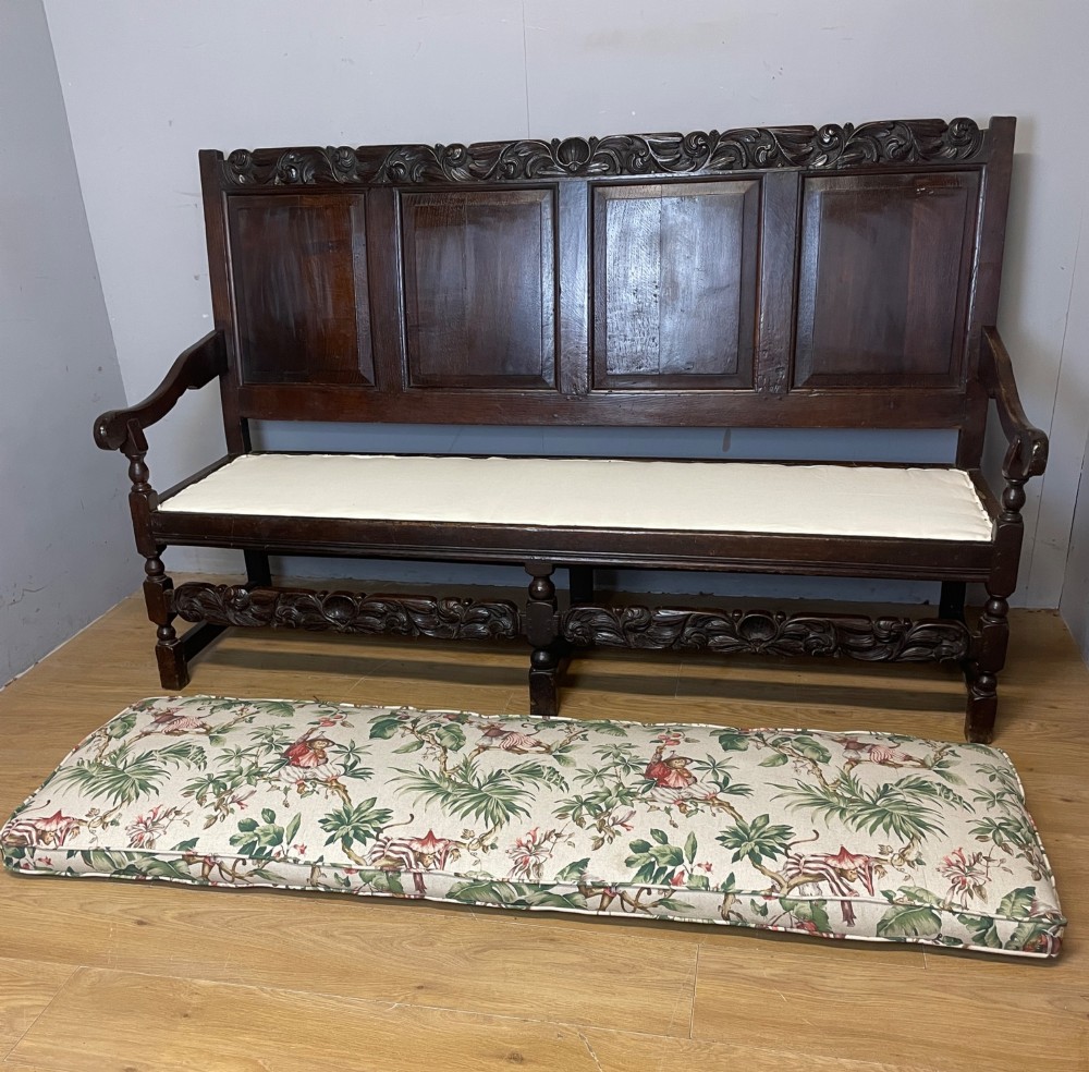 An antique dark oak wooden settle featuring four raised back panels, detailed floral relief carving along the crest and apron, turned legs, and a floral pattern cushion. View XIII