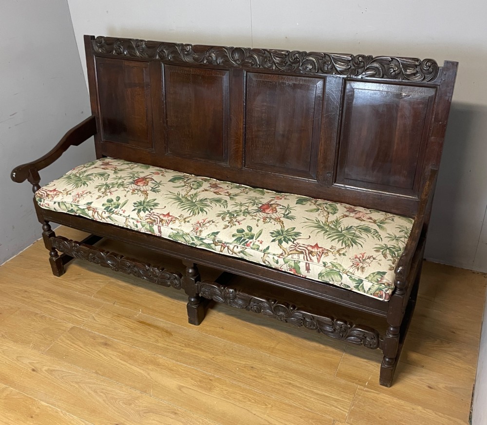An antique dark oak wooden settle featuring four raised back panels, detailed floral relief carving along the crest and apron, turned legs, and a floral pattern cushion. View XV