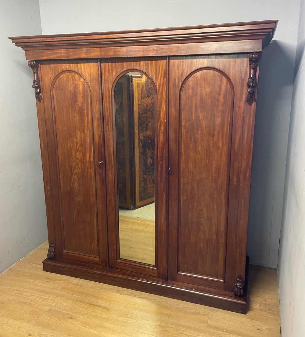 A 19th-century flame mahogany triple wardrobe with an arched central mirrored door flanked by two panelled doors, topped with a stepped cornice and carved corbels.