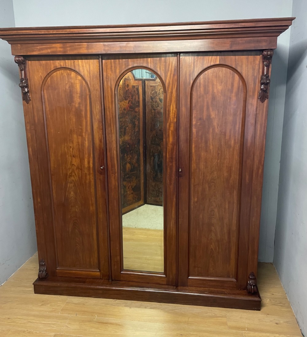 A 19th-century flame mahogany triple wardrobe with an arched central mirrored door flanked by two panelled doors, topped with a stepped cornice and carved corbels. View II