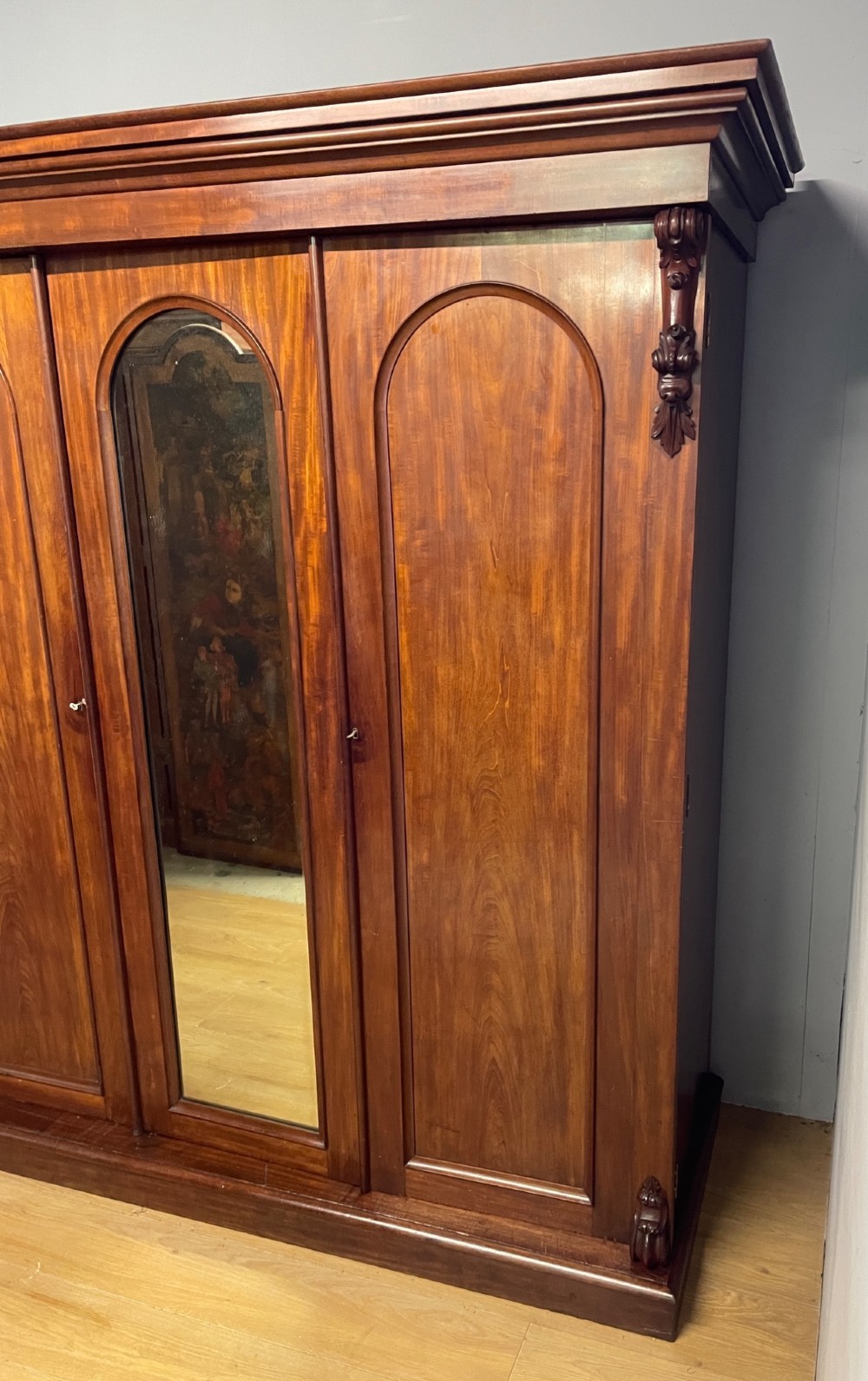 A 19th-century flame mahogany triple wardrobe with an arched central mirrored door flanked by two panelled doors, topped with a stepped cornice and carved corbels. View IV