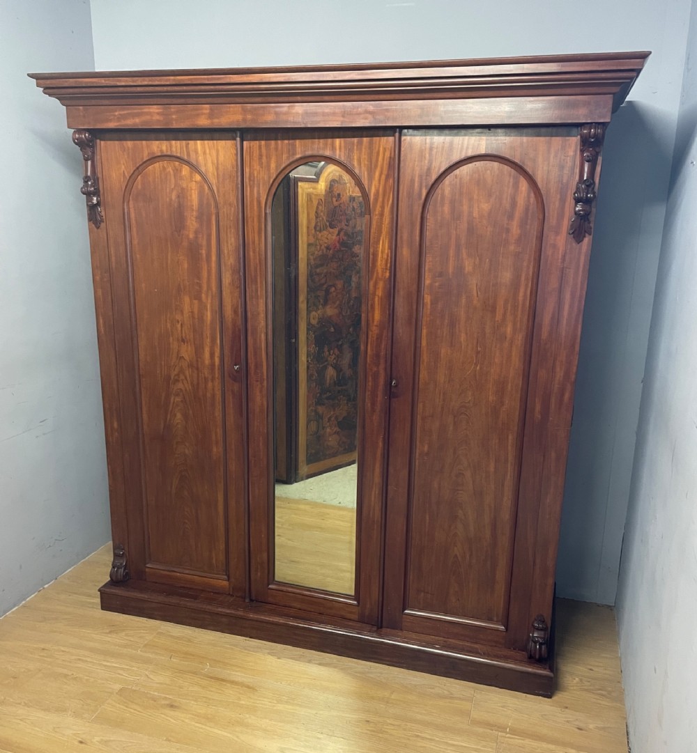 A 19th-century flame mahogany triple wardrobe with an arched central mirrored door flanked by two panelled doors, topped with a stepped cornice and carved corbels. View X