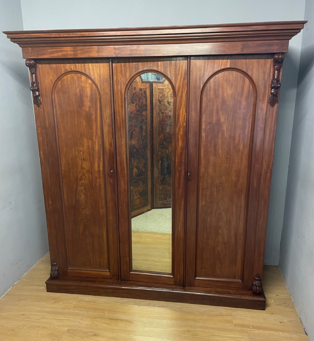 A 19th-century flame mahogany triple wardrobe with an arched central mirrored door flanked by two panelled doors, topped with a stepped cornice and carved corbels. View XI