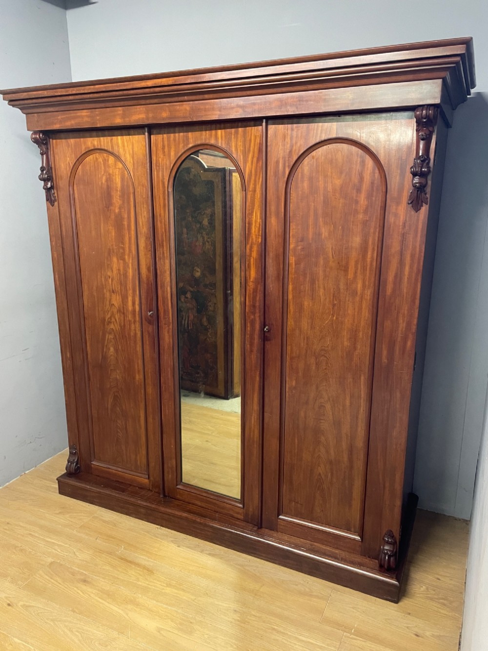 A 19th-century flame mahogany triple wardrobe with an arched central mirrored door flanked by two panelled doors, topped with a stepped cornice and carved corbels. View XII