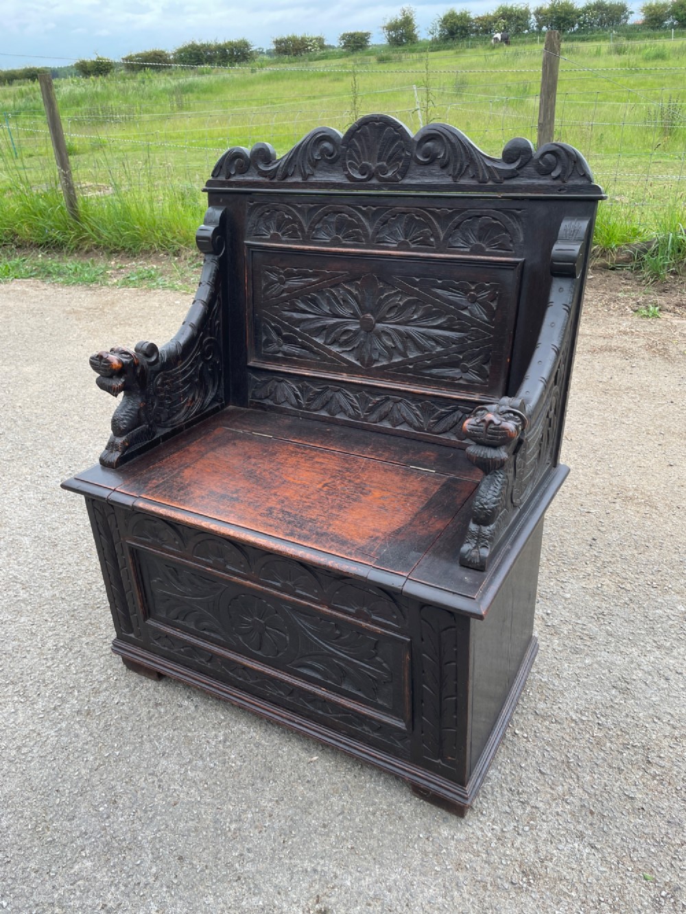 A 19th-century dark oak box settle with carved floral panels, lion mask terminals, a decorative cresting rail, and a lift-up storage seat.