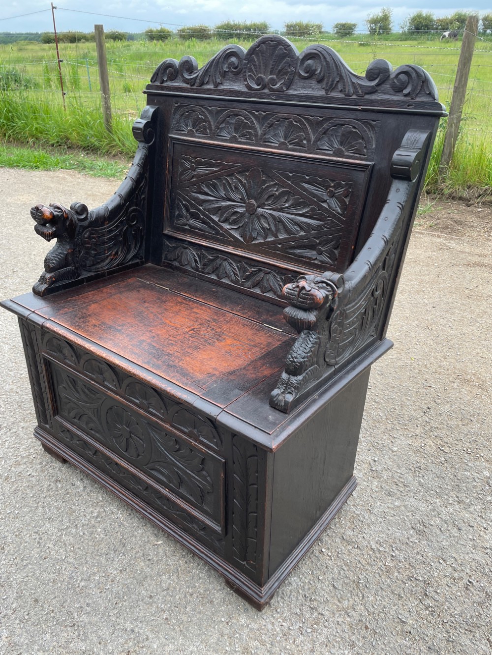 A 19th-century dark oak box settle with carved floral panels, lion mask terminals, a decorative cresting rail, and a lift-up storage seat. View II