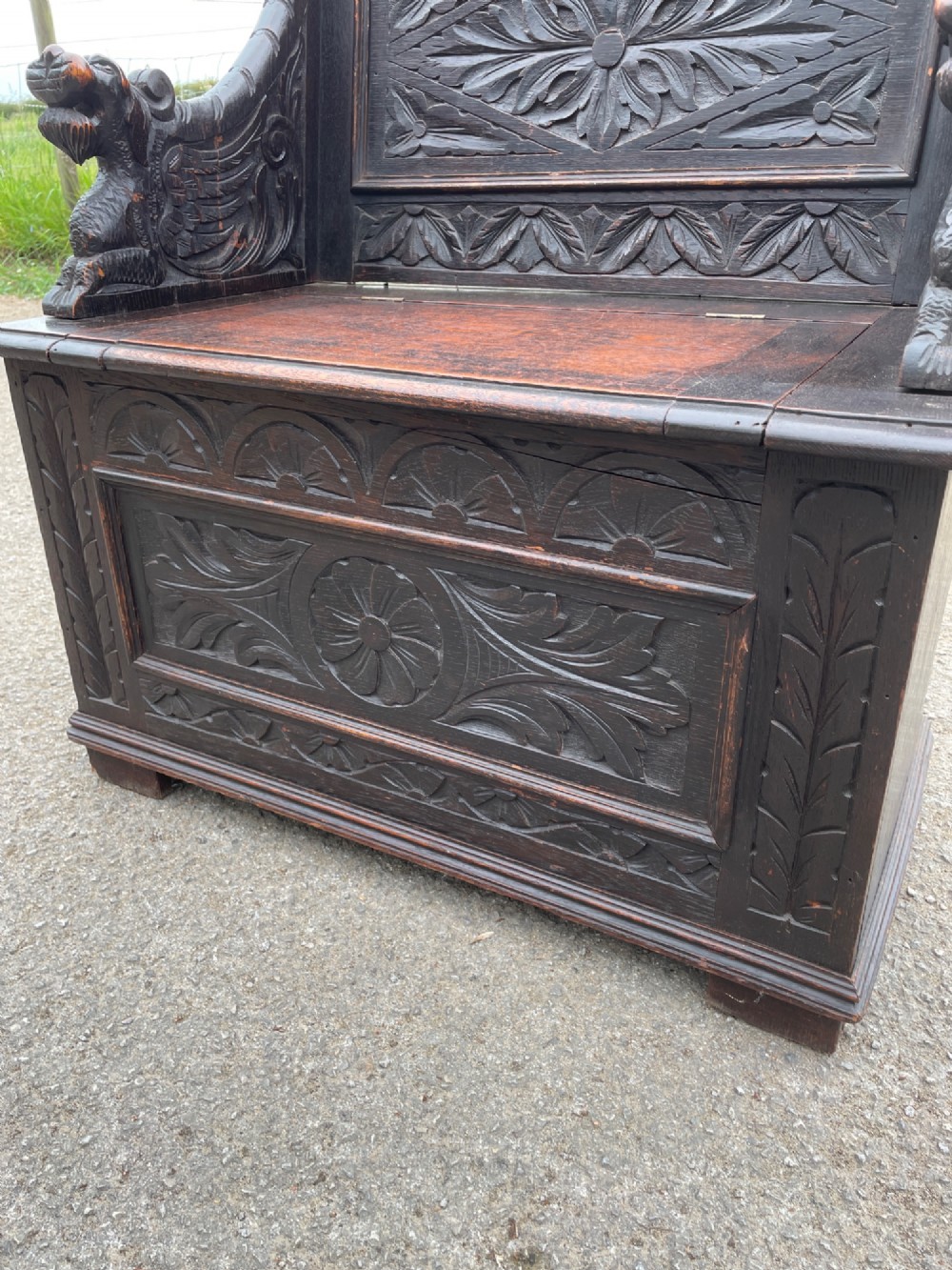 A 19th-century dark oak box settle with carved floral panels, lion mask terminals, a decorative cresting rail, and a lift-up storage seat. View III