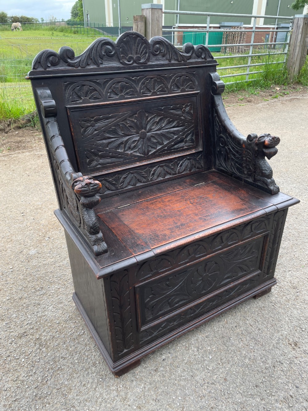 A 19th-century dark oak box settle with carved floral panels, lion mask terminals, a decorative cresting rail, and a lift-up storage seat. View IV