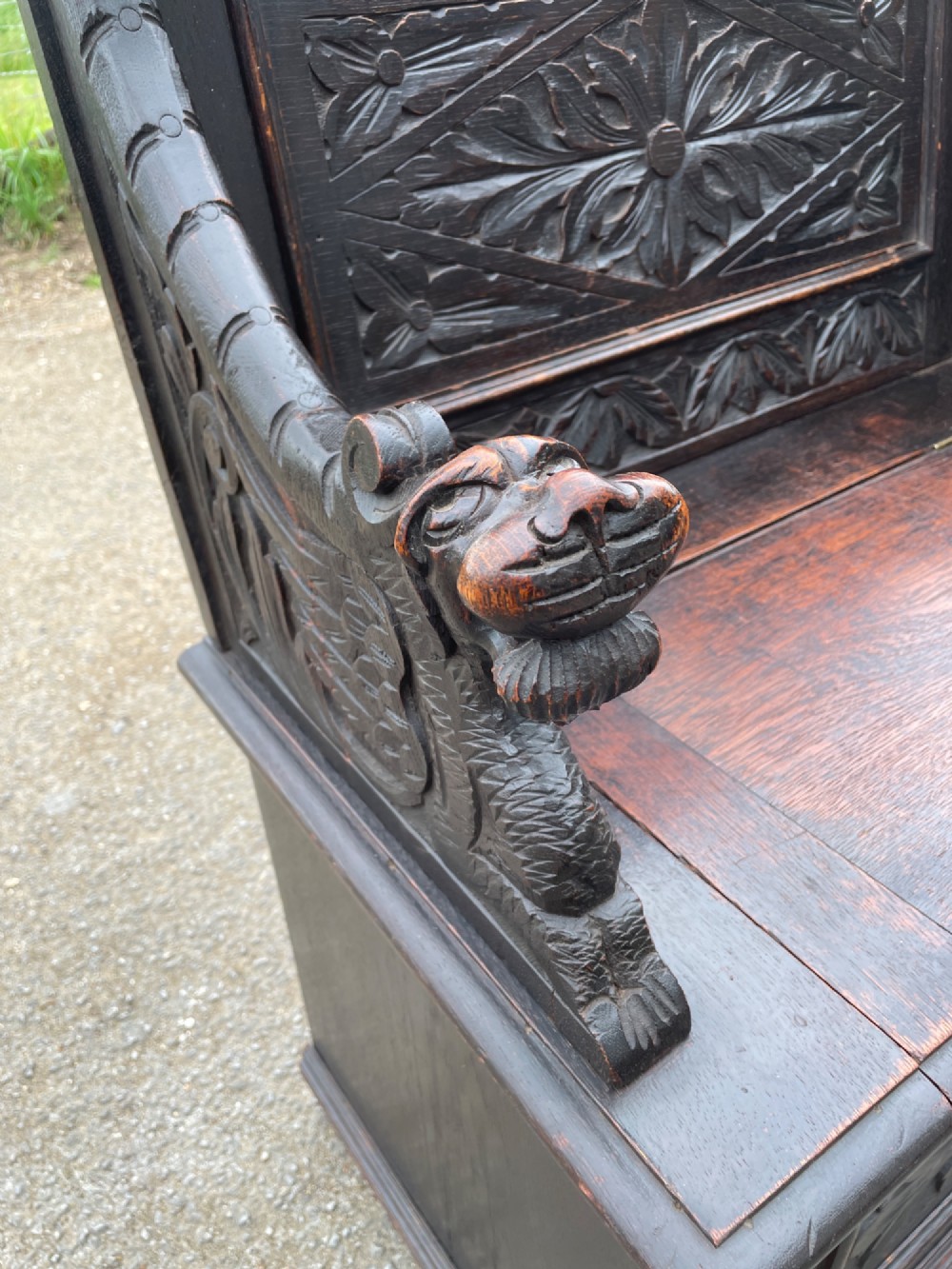 A 19th-century dark oak box settle with carved floral panels, lion mask terminals, a decorative cresting rail, and a lift-up storage seat. View V