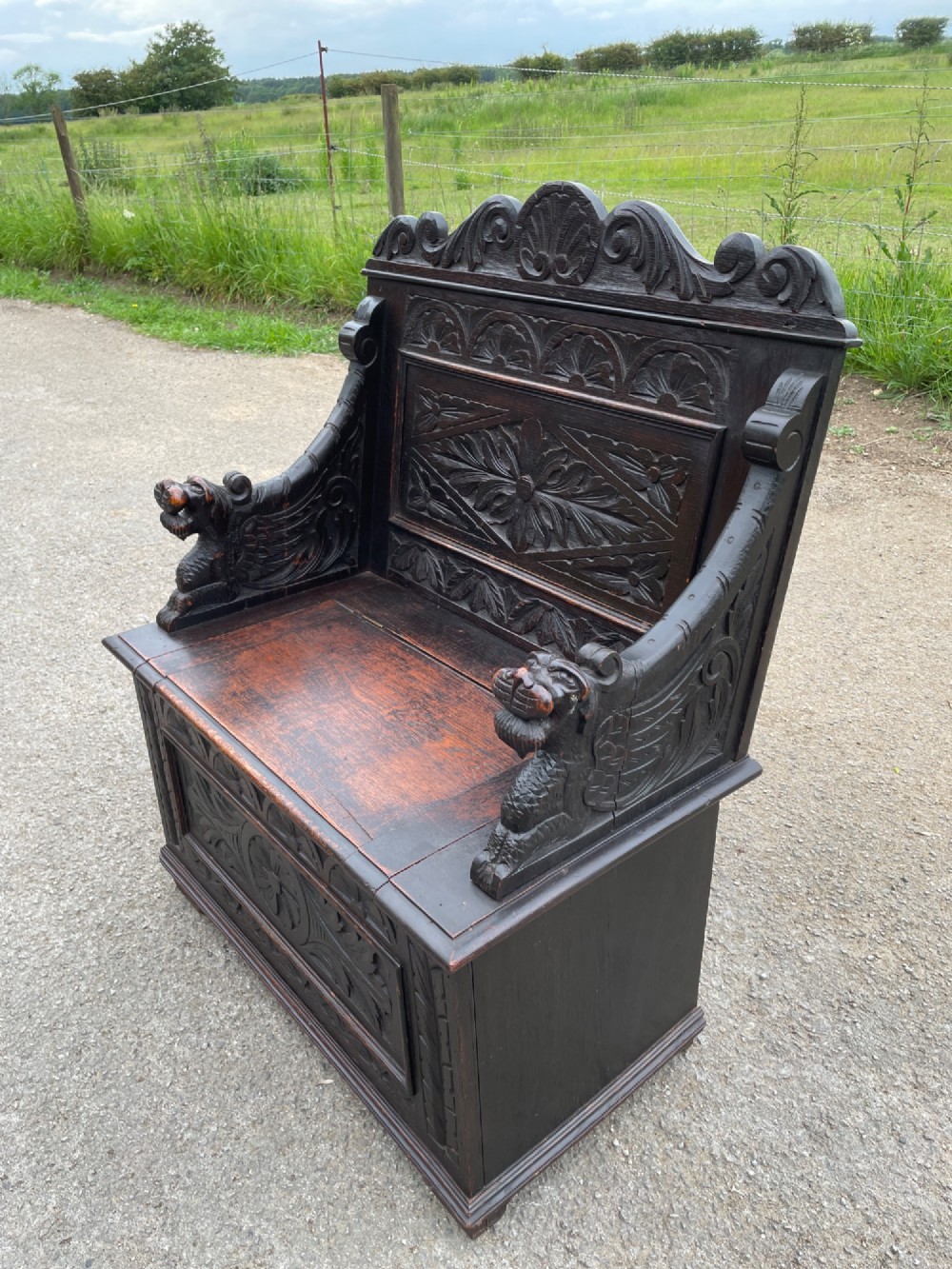 A 19th-century dark oak box settle with carved floral panels, lion mask terminals, a decorative cresting rail, and a lift-up storage seat. View VI