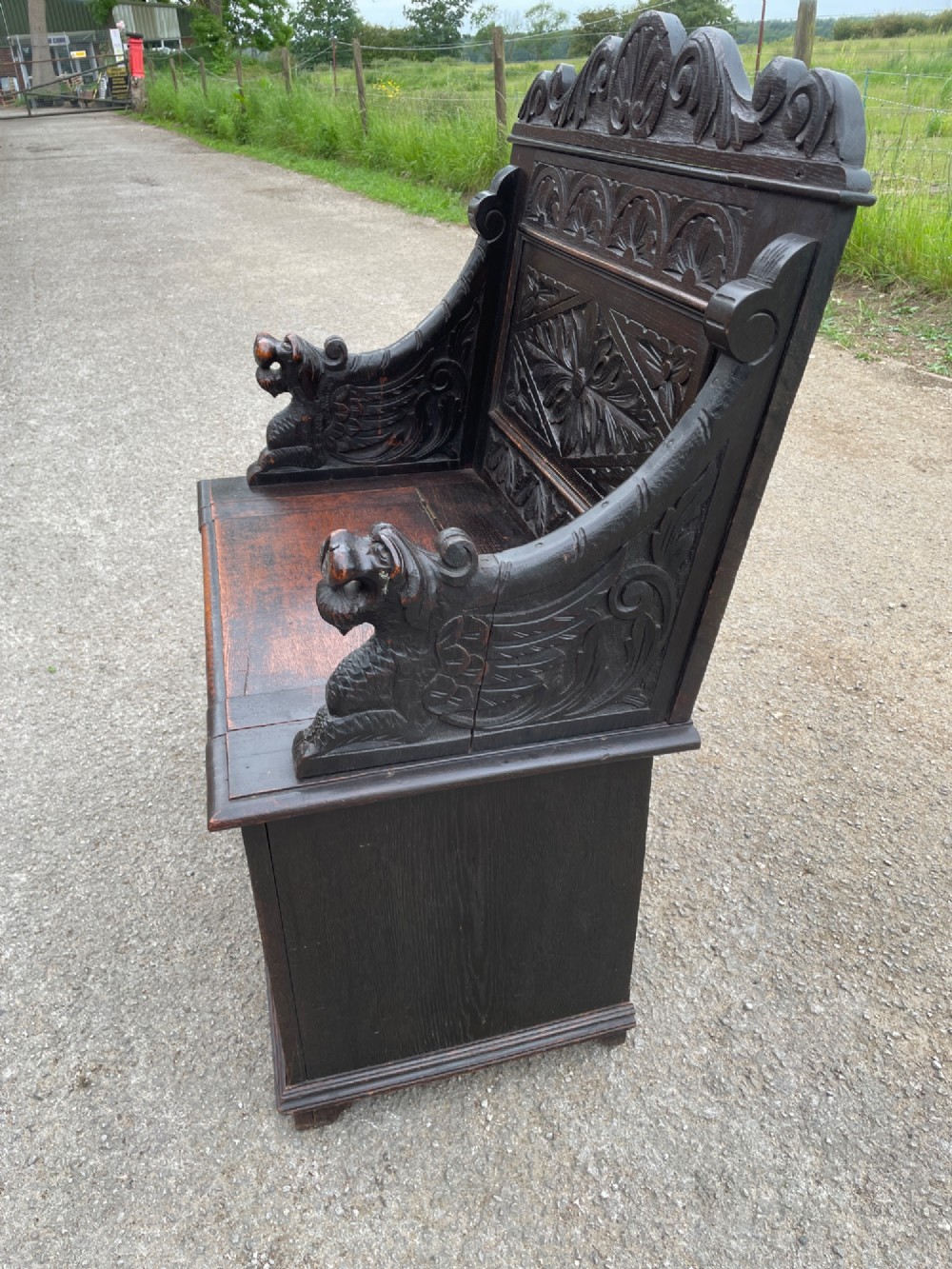 A 19th-century dark oak box settle with carved floral panels, lion mask terminals, a decorative cresting rail, and a lift-up storage seat. View VII