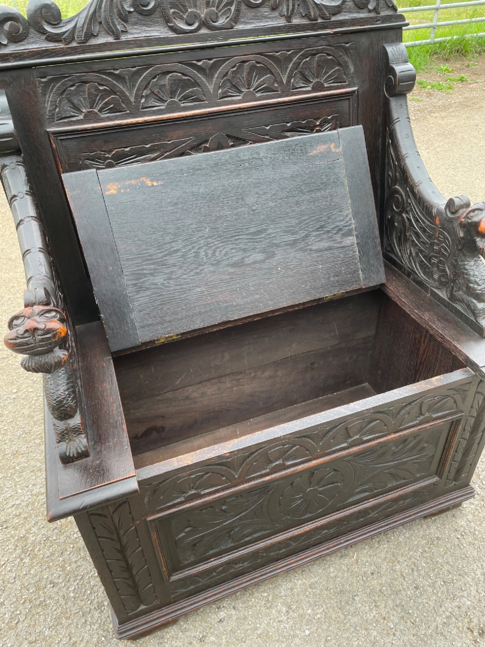A 19th-century dark oak box settle with carved floral panels, lion mask terminals, a decorative cresting rail, and a lift-up storage seat. View IX