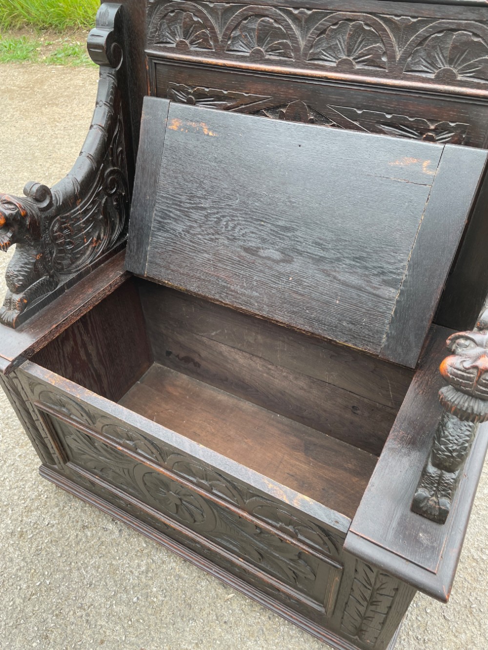 A 19th-century dark oak box settle with carved floral panels, lion mask terminals, a decorative cresting rail, and a lift-up storage seat. View X